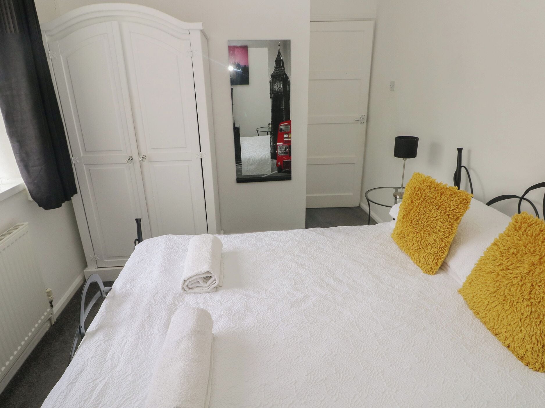 A neatly made bed with a rolled towel, a mirror reflecting a clock, and a red bus outside a door.