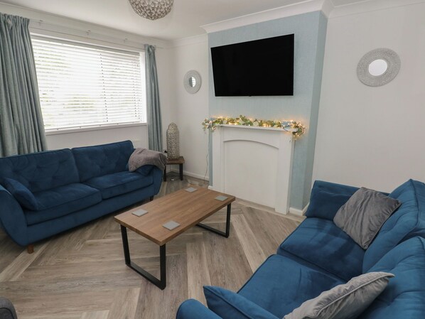 A living room with a blue sofa, a wooden coffee table, a fireplace, and a flat-screen TV mounted on the wall. - Cwtch Ar Y Mor (Burry Port)