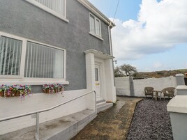 A residential building with a grey exterior, white window frames, and a white door. There are flower boxes with pink and purple flowers on the ground floor. A gravel path leads to a seating area with wicker chairs and a table.
