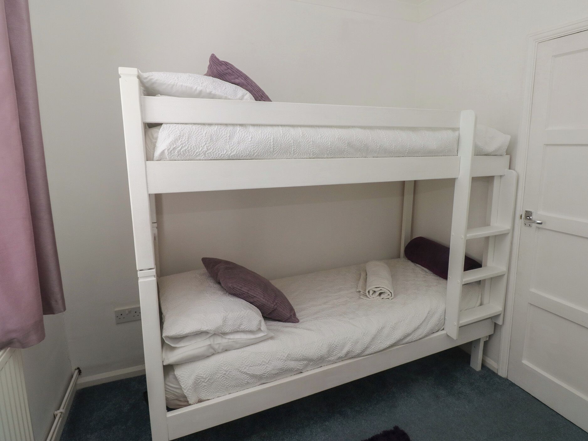 A bunk bed with white bedding and purple pillows, a white door, and a radiator.