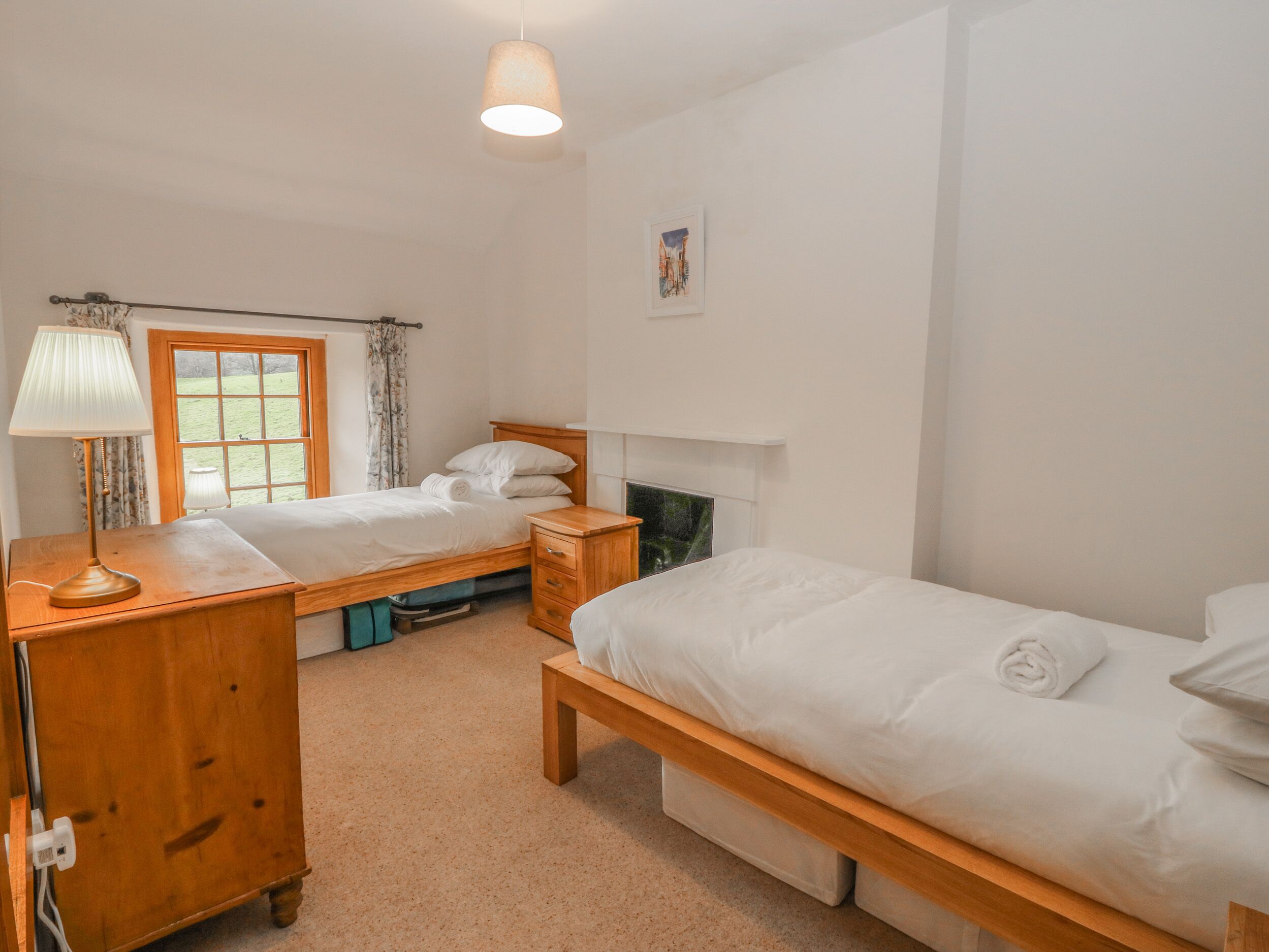 A room with two single beds, a wooden nightstand with a lamp, a window with curtains, and a painting on the wall.