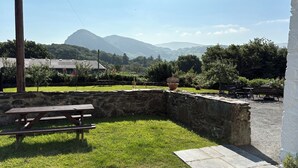 Un muro de piedra, una mesa de picnic y un banco en un espacio verde con montañas al fondo.