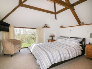 A bedroom with a bed, a chair, a nightstand, a TV, and a window with a view of trees. - Sleepy Fox (Bodmin)