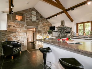 A modern kitchen with a stone wall, wooden beams, a large island with bar stools, and a stone fireplace. - Sleepy Fox (Bodmin)