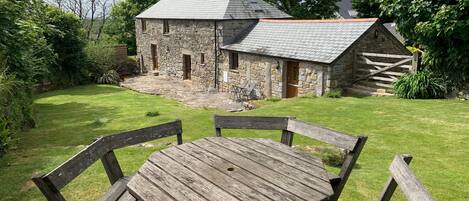 A stone building with a sloped roof, a wooden table with benches, and a grassy yard.