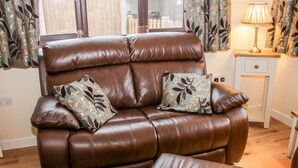 A brown leather sofa with two patterned cushions, a matching ottoman, and a bedside table with a lamp.