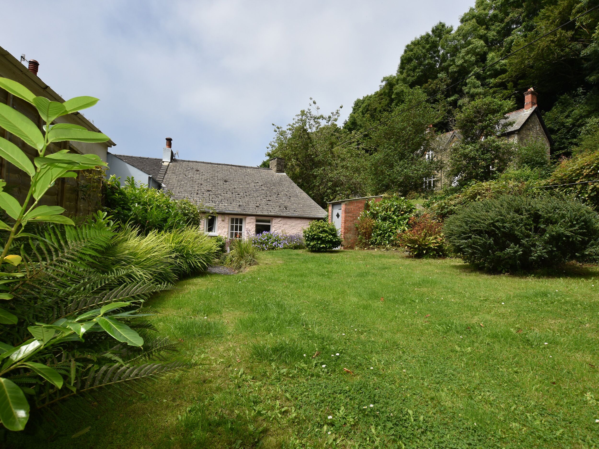 A traditional stone house with a sloped roof, surrounded by lush greenery and a well-maintained lawn.