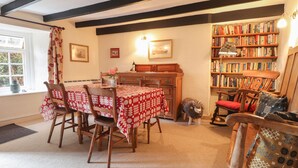 Un coin repas accueillant, avec une table dressée pour quatre personnes, entourée de chaises en bois. Une bibliothèque remplie de livres se trouve à droite, et on peut apercevoir un petit chien près de la bibliothèque.