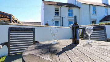 A wooden table with two wine glasses and a bottle, set against a white building with multiple windows.