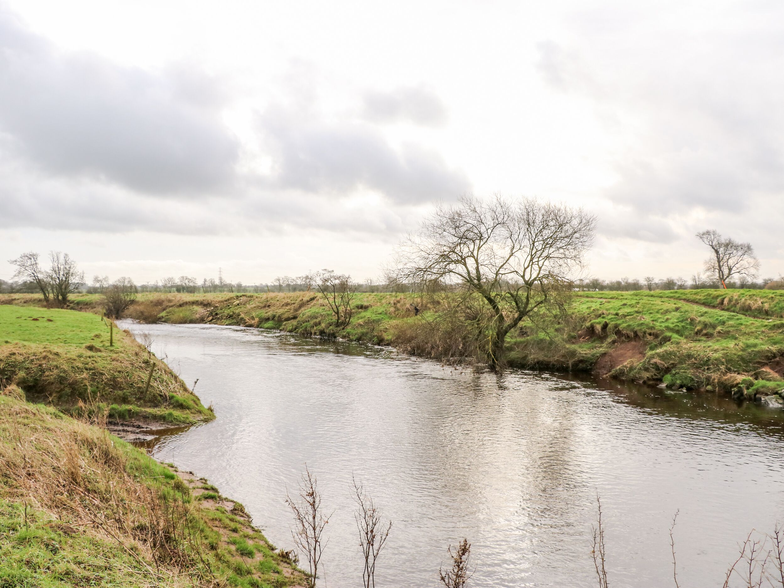 A river flowing through a rural landscape with grassy banks and leafless trees.