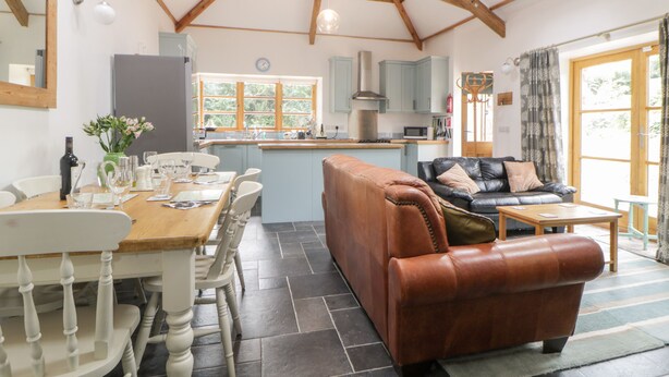 A spacious kitchen with a central island, a dining area with a wooden table and chairs, a leather sofa, and a large window providing natural light. - The Old Haybarn (Newton Abbot)