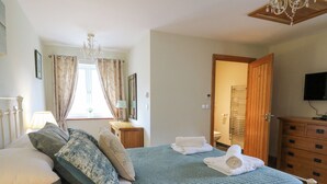 A bedroom with a bed, pillows, a bedside table with a lamp, a wooden dresser, a television, and a bathroom visible through an open door. - Yr Hen Beudy (Aberystwyth)