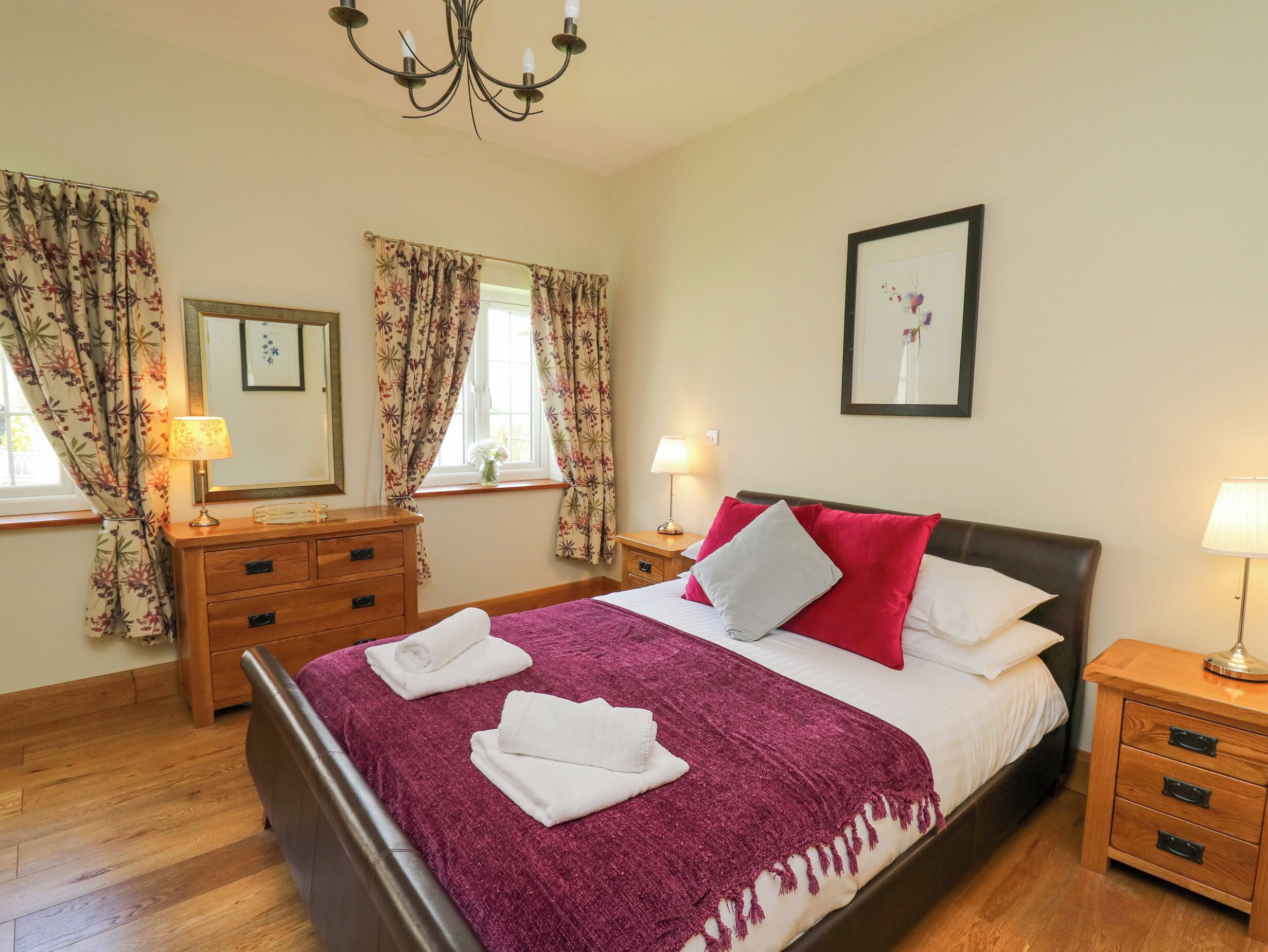 A neatly arranged bedroom with a bed, two bedside tables, a dresser, a chandelier, and a framed picture on the wall.
