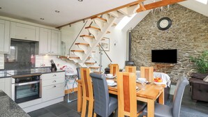 A modern kitchen with white cabinets, a built-in oven, and a dining area with a wooden table and chairs. A stone wall and a television are visible in the background. - Yr Hen Beudy (Aberystwyth)