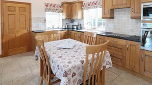A kitchen with wooden cabinets, a dining table with a patterned tablecloth, and a tiled backsplash. - Bryn Eiddon (Pwllheli)