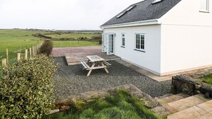 A white house with a sloped roof, a gravel patio, and a wooden table. - Bryn Eiddon (Pwllheli)