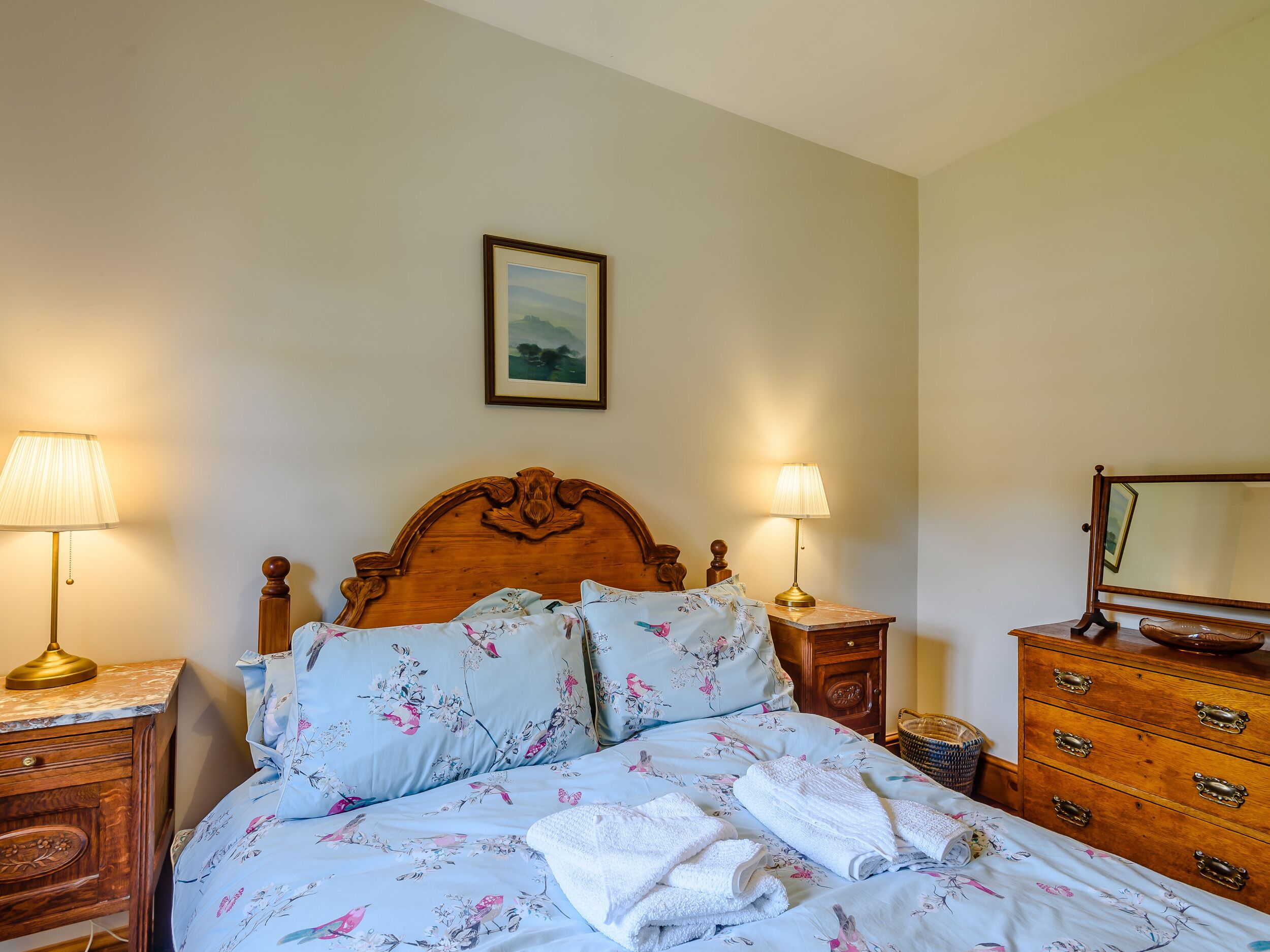 A neatly made bed with floral bedding, two bedside lamps, a wooden headboard, a small wooden nightstand, a dresser with drawers, and a framed picture on the wall.