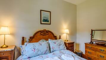 A neatly made bed with floral bedding, two bedside lamps, a wooden headboard, a small wooden nightstand, a dresser with drawers, and a framed picture on the wall.