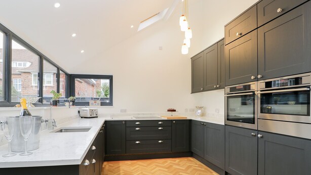 Une cuisine moderne avec des meubles de couleur sombre, un parquet en bois disposé en chevrons, une grande fenêtre et des appareils électroménagers intégrés.