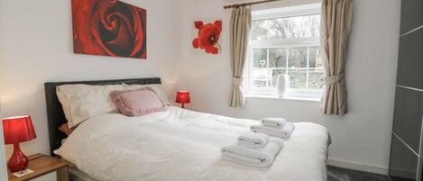 Un lit bien fait, avec du linge de lit blanc et des coussins roses, une table de chevet avec une lampe rouge, une fenêtre avec des rideaux et un tableau accroché au mur.