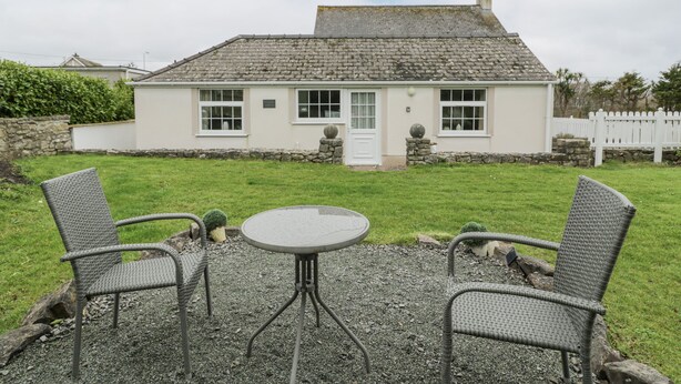 A house with a white exterior, a stone chimney, and a gravel patio with two wicker chairs and a round table. - Halcyon Annexe (Moelfre)