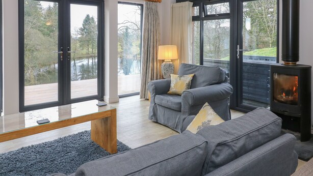 A modern living room with a grey sofa, a wooden coffee table, and a black wood-burning stove. - Tarn Lodge (Windermere)