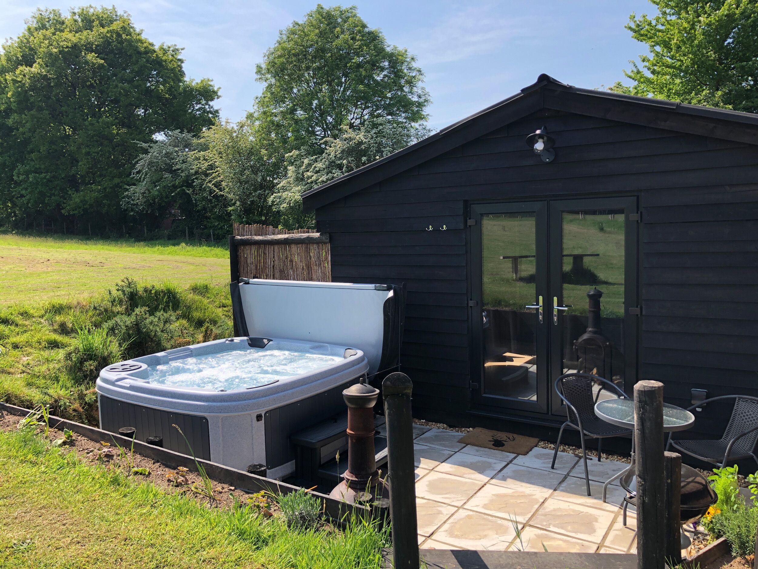 Een bubbelbad naast een houten chalet met een glazen deur, een terras met stoelen en een gazon.
