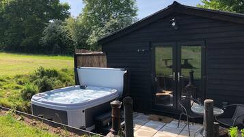 Een bubbelbad naast een houten chalet met een glazen deur, een terras met stoelen en een gazon.