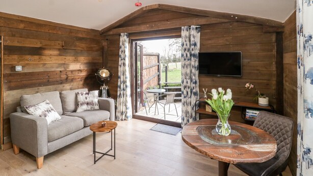 A cozy living room with a sofa, a coffee table, a TV mounted on the wall, and a wooden ceiling. - Deer Lodge (Wimborne)