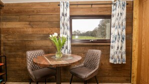 A wooden interior with a round table, two cushioned chairs, a vase with white flowers, and a window with curtains. - Deer Lodge (Wimborne)