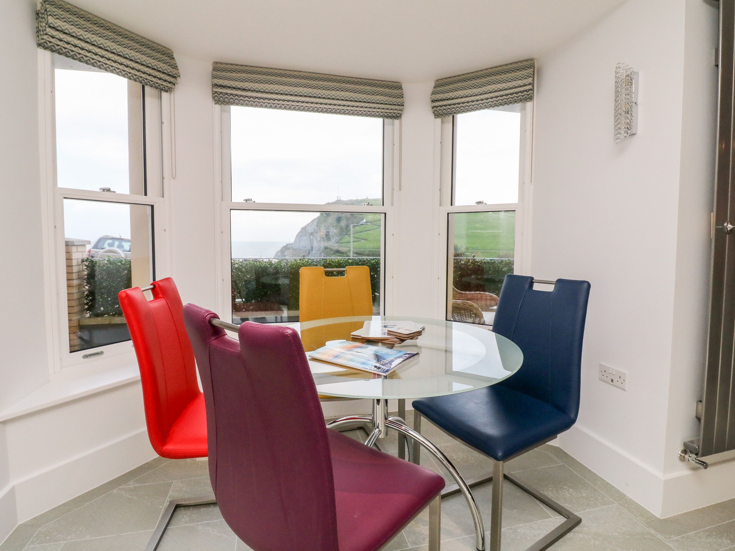 A dining area with a round glass table, four chairs, and large windows with curtains.