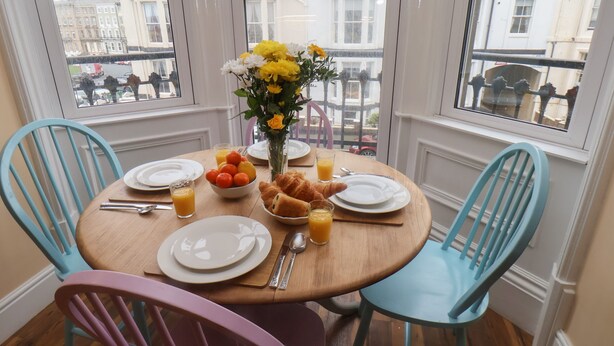 A breakfast table set with plates, a bowl of fruit, croissants, and a vase of flowers. - Apartment 4 (Whitby)