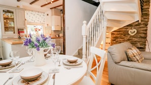 A dining area with a round table set for four, white chairs, and a vase of flowers. A staircase with a white railing leads to a living area with a sofa and a heart-shaped decoration on the wall. - Dalesway Cottage (Windermere)