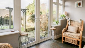 A cozy room with a wicker chair, a small table, and a view of a garden through large glass doors. - Dalesway Cottage (Windermere)