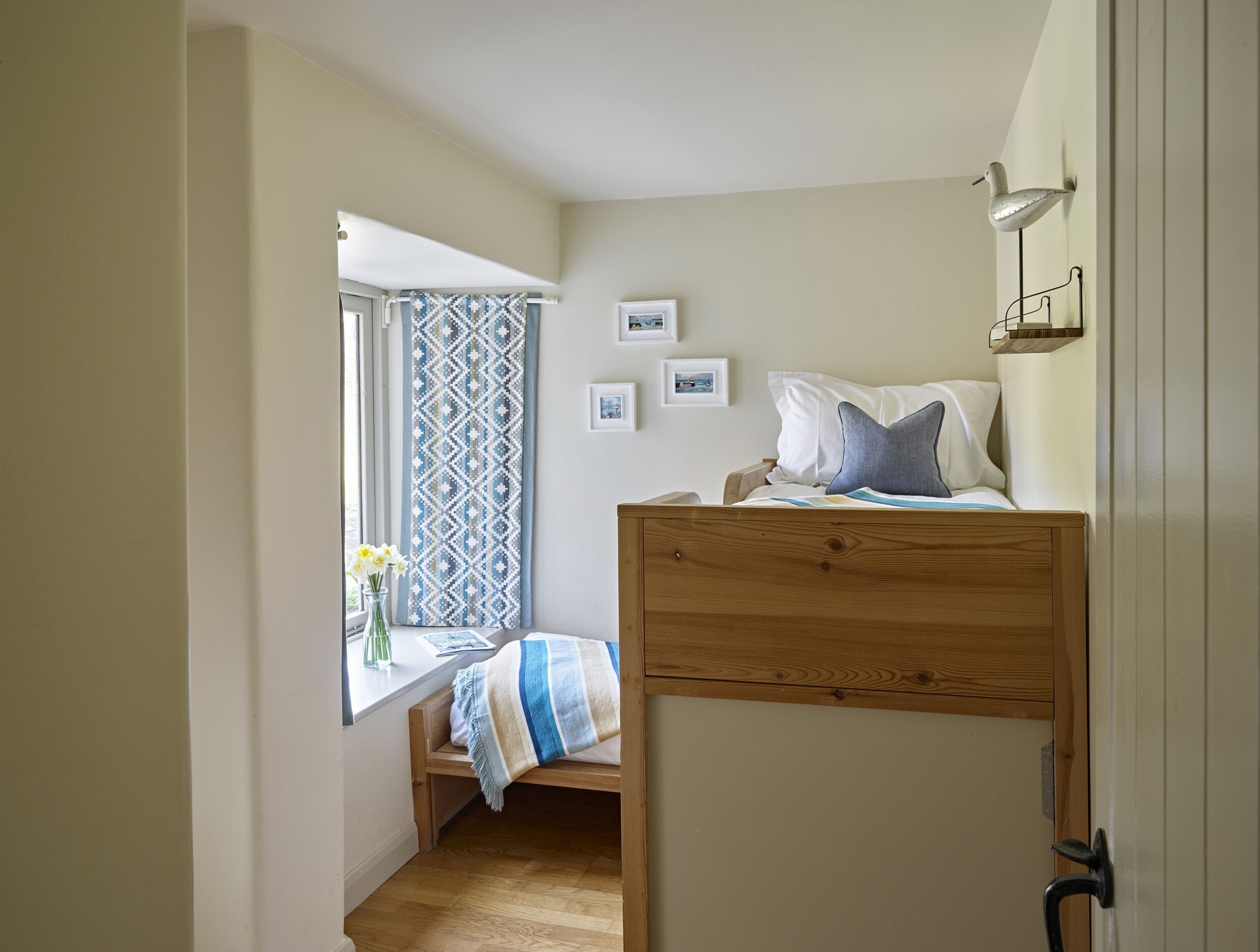 A small, cozy bedroom with a wooden bed, a window with patterned curtains, and a bedside table with a vase of flowers.
