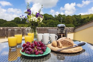 Una mesa de desayuno con hogaza de pan, un cuchillo, un plato de uvas, vasos de jugo de naranja y un florero con flores.