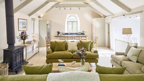 A spacious loft with high ceilings, exposed wooden beams, a cozy green sofa, a fireplace, and a kitchen in the background. - Garden House (Totnes)