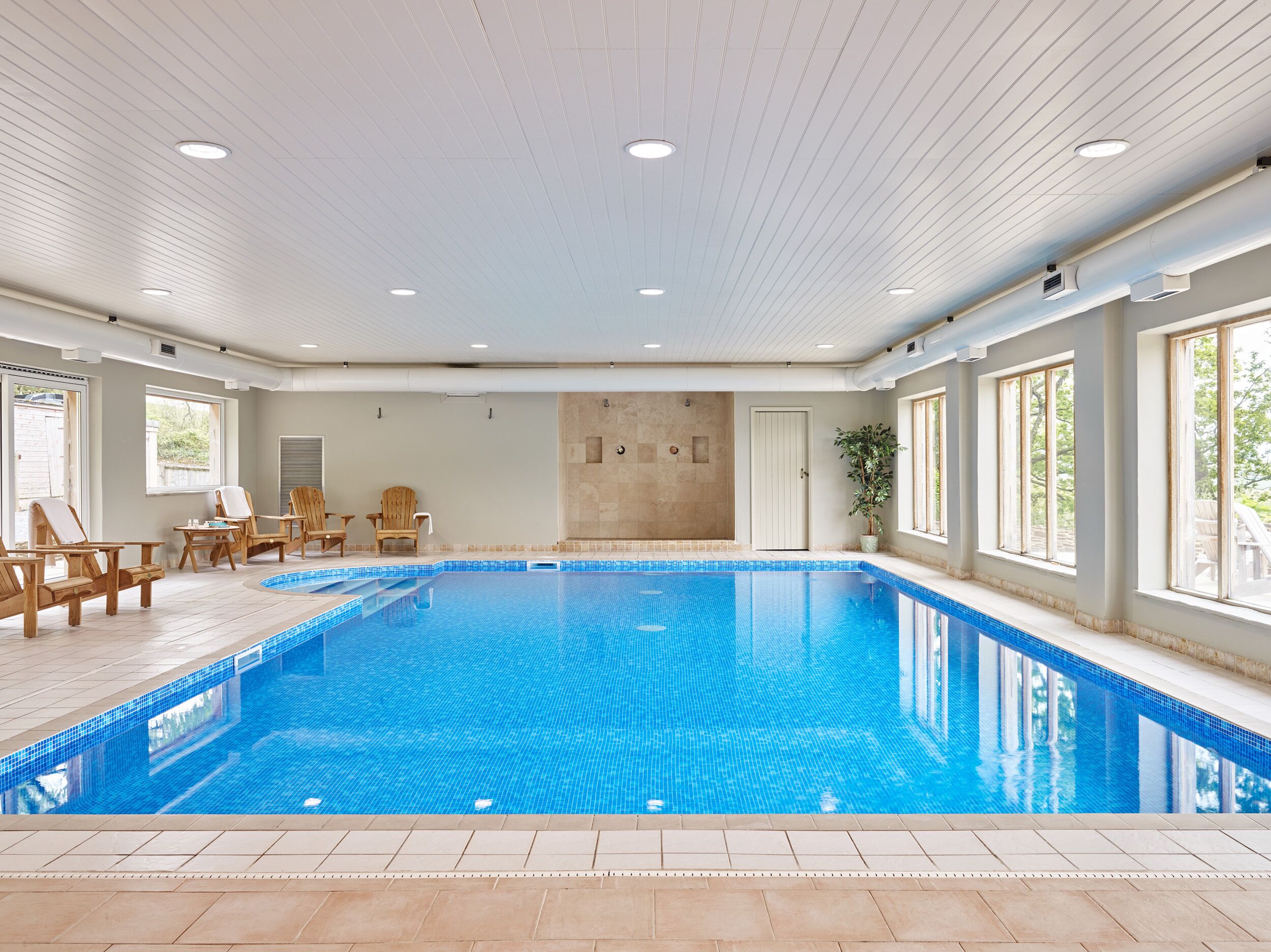 A large indoor swimming pool with clear blue water, surrounded by wooden chairs and a potted plant.