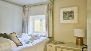 A neatly made bed with folded towels, a bedside lamp, a framed picture, and a window with curtains.