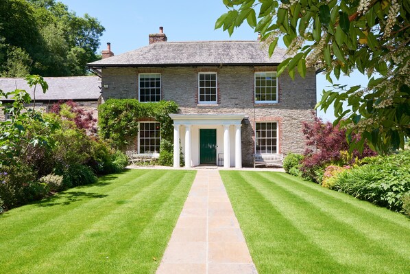 A stone building with a central entrance flanked by columns, surrounded by well-maintained lawns and various shrubs. - Gitcombe House (Totnes)