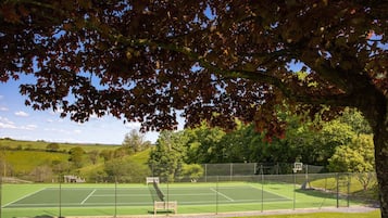 An outdoor tennis court with a net, surrounded by a fence, and a bench in the foreground.
