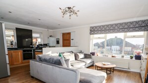 A modern living room with a grey sofa, wooden flooring, a kitchen in the background, and a large window with a view of houses. - Honeybees Haven (Hunstanton)