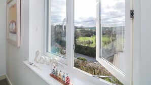 Ein Zimmer mit großem Fenster und Blick auf eine grüne Landschaft, einem Regal mit Dekoration und einem gerahmten Bild an der Wand.