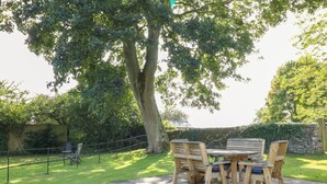An outdoor patio with a stone table and wooden chairs, surrounded by a stone wall and a grassy area with trees. - Holmefield (Bakewell)