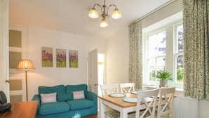 A cozy living room with a teal sofa, a wooden dining table with white chairs, a floor lamp, and a window with patterned curtains. - Holmefield (Bakewell)