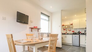 A dining area with a wooden table and chairs, a TV mounted on the wall, and a kitchen in the background. - Y Bwthyn, Newport (Newport)