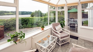 A conservatory with white wicker furniture, a glass table, and a view of a river and greenery.