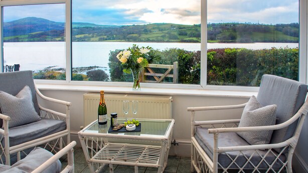 A room with two wicker chairs, a glass table, and a view of a lake and hills.