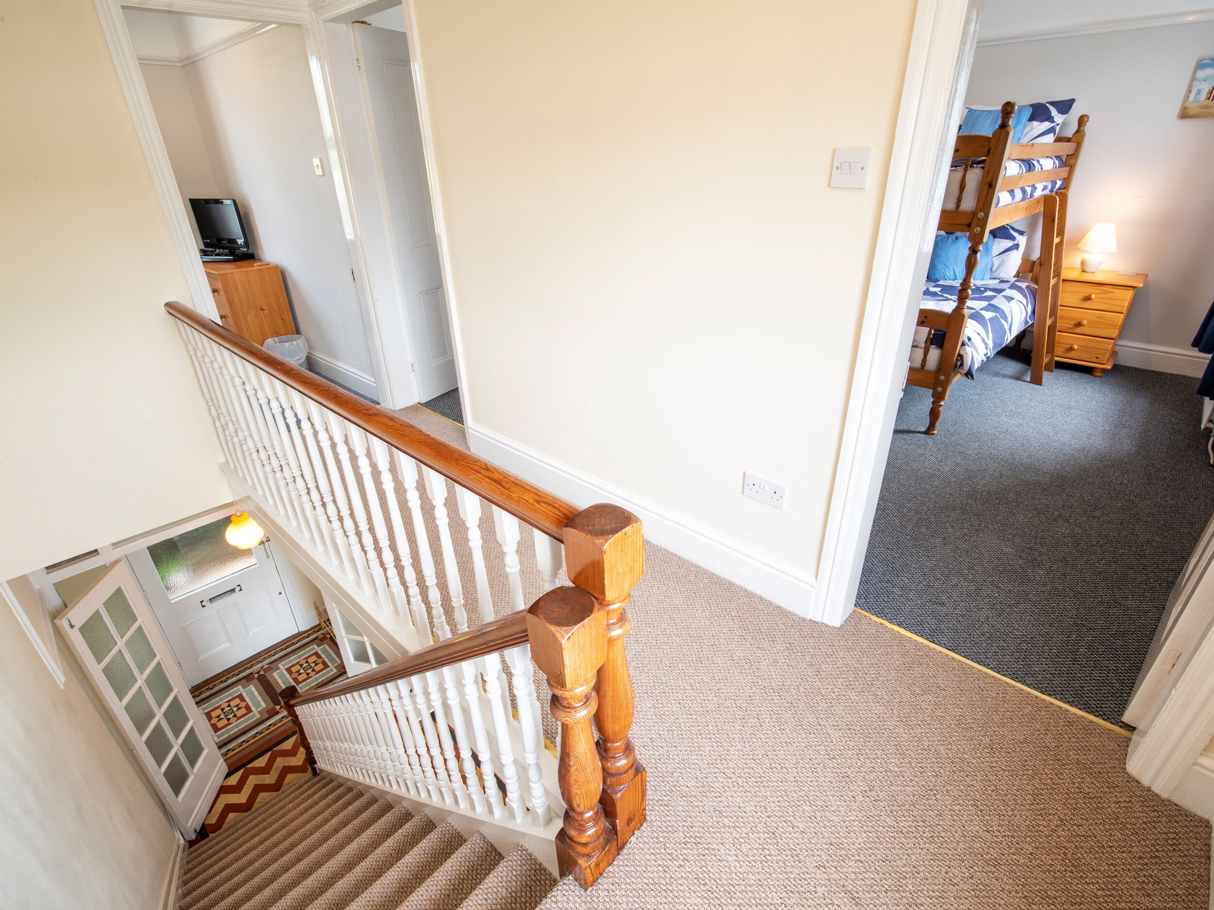 A staircase with a wooden handrail and white balusters, carpeted steps, a door with frosted glass panels, and a bedroom with a bunk bed and a bedside table with a lamp.