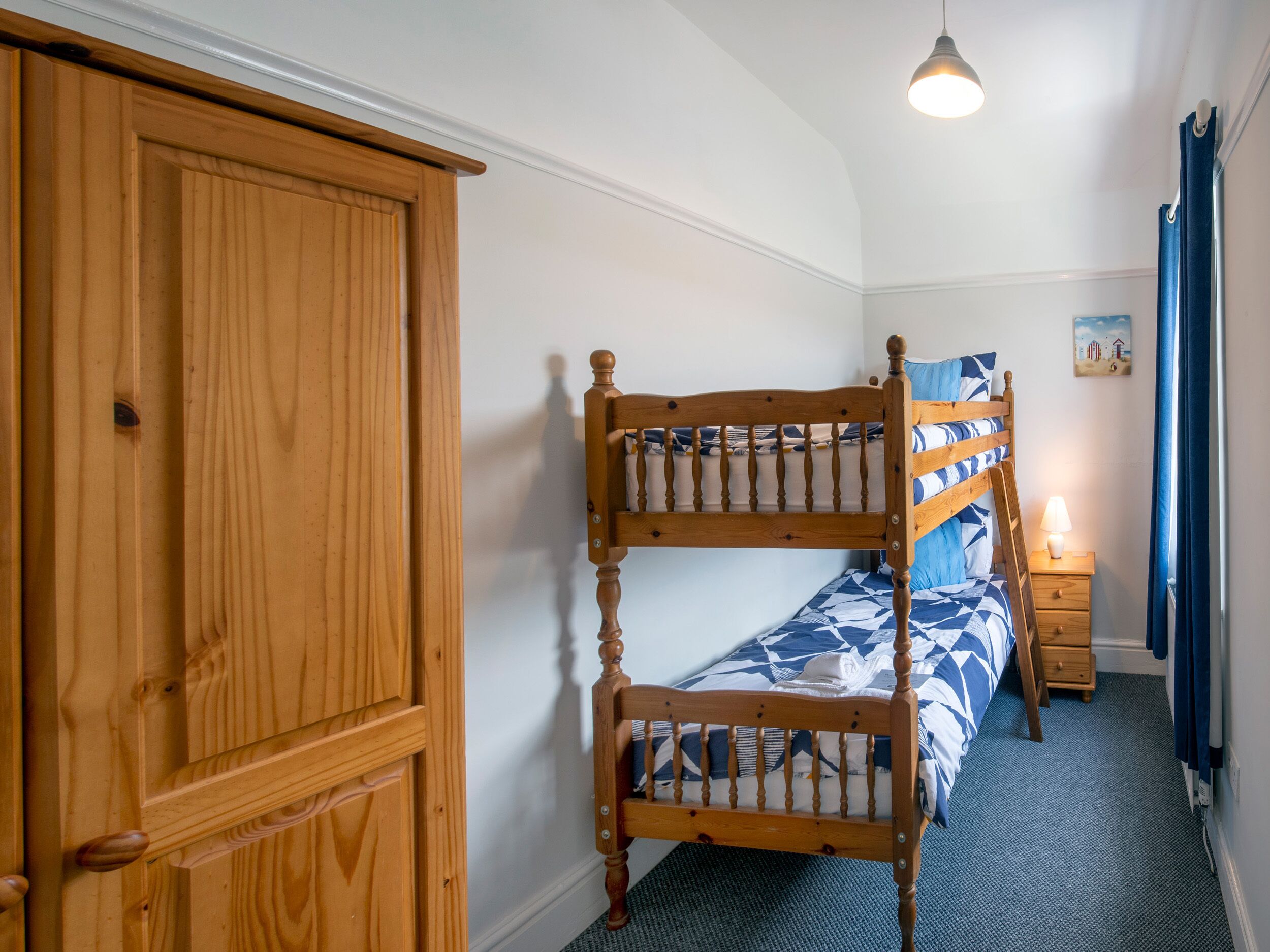 A bedroom with a bunk bed, a wooden wardrobe, a bedside table with a lamp, and a small picture on the wall.
